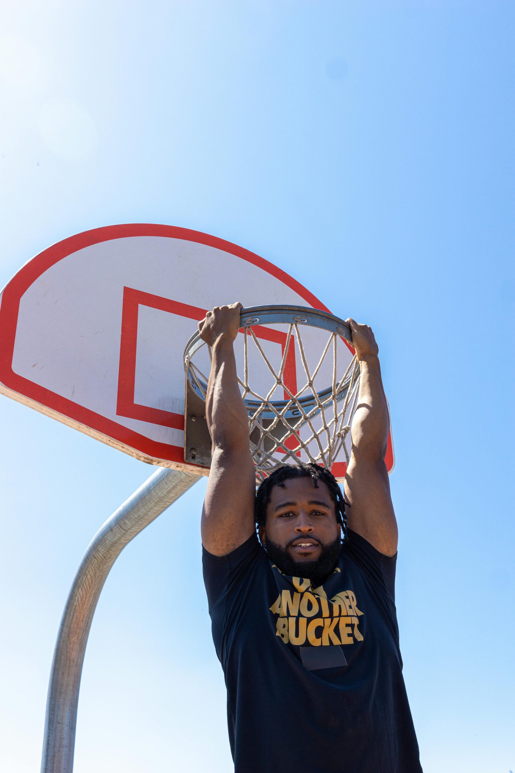 A person hangs from a basketball hoop on a sunny day, enjoying a playful moment on the court.